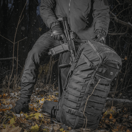 Person med M-Tac Rygsæk Gen.II Elite Large i skov og våben til transport, slidstærk Cordura materiale, ergonomisk design.