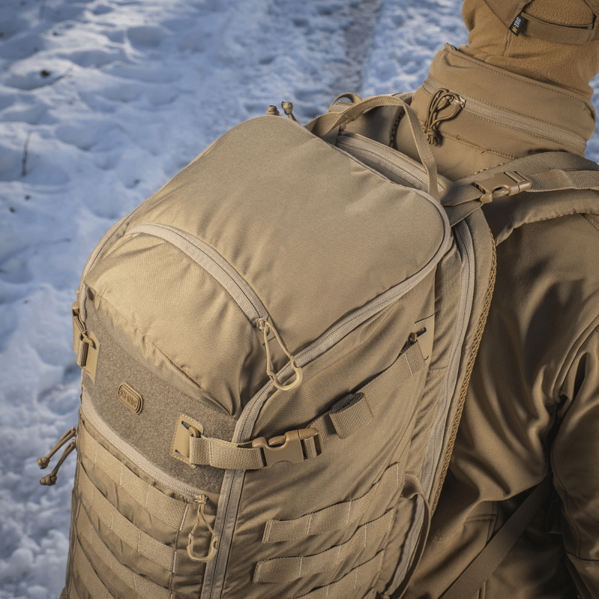 M-Tac Rygsæk Gen.II Elite Large bæres i sneklædt landskab, viser robusthed og design i Cordura 330D materiale, velegnet til våbentransport.