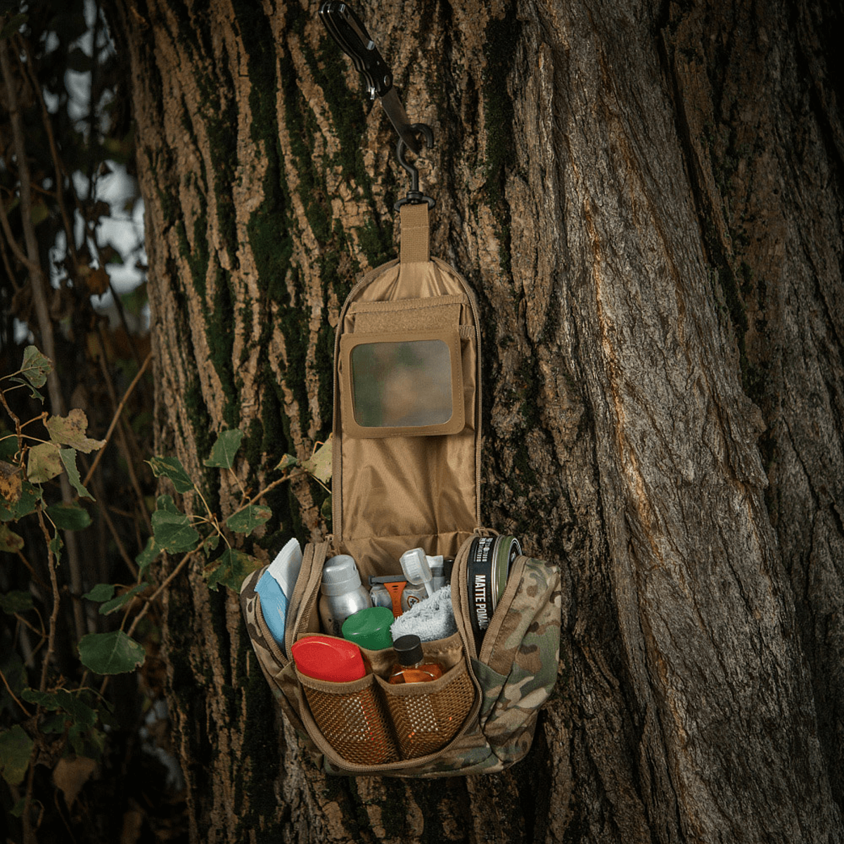 M-Tac Toilettaske Elite Gen.II hænger åben på et træ fyldt med hygiejneartikler, perfekt organiseret til rejsebrug.