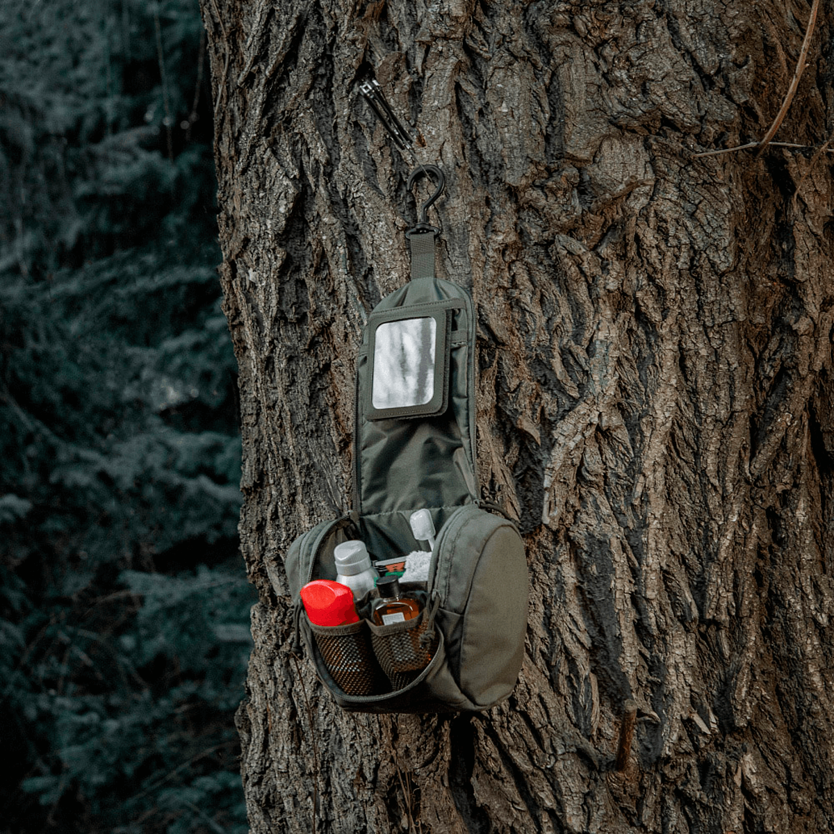 M-Tac Toilettaske Elite Gen.II hængende på et træ, fyldt med hygiejneartikler som tandpasta, barbermaskine og lotion.
