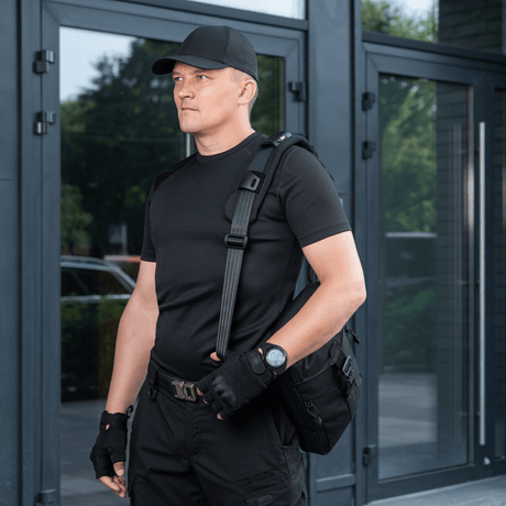 Man carrying a bag with M‑Tac Shoulder Strap Pad 50 mm Elite for increased comfort and support, wearing black clothing in front of a modern building.