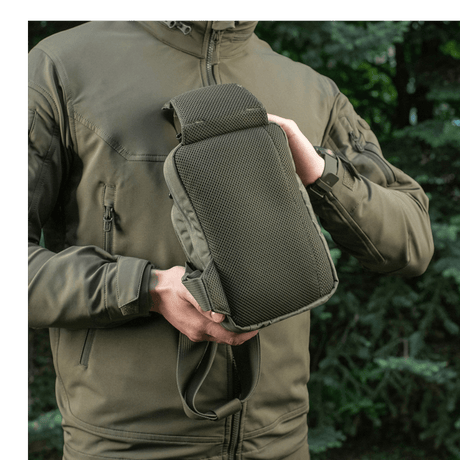 Person showing M-Tac Sling Pistol Bag Elite Hex outdoors in a woodland setting, wearing a zippered jacket.