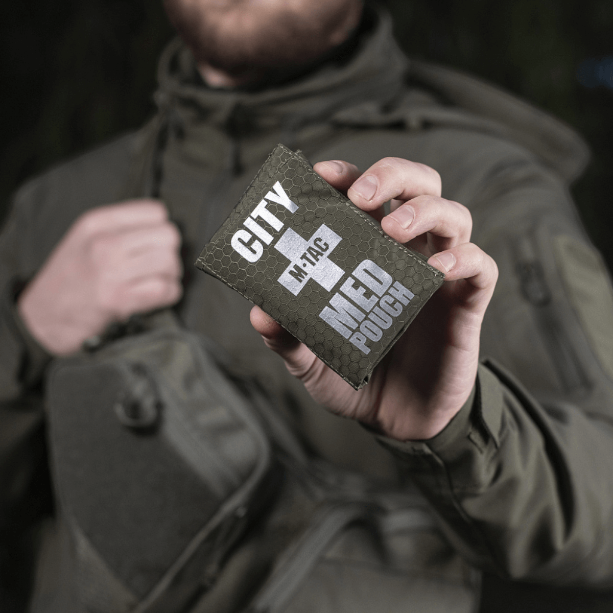 M-Tac City Med Pouch Hex holdt af person iført jakke, kompakt førstehjælpskit i Hex Polyester med Velcro lukning, M-Tac logo vises.