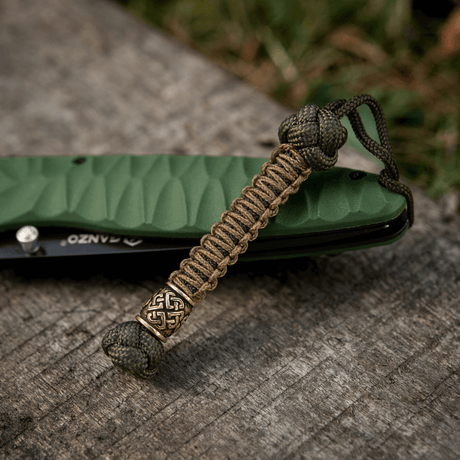M-Tac Knife Lanyard Celtic in braided paracord next to a green pocketknife on a wood surface.