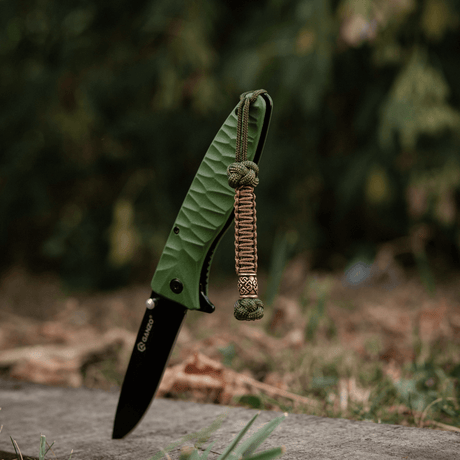 M-Tac Knife Lanyard Celtic - green folding knife with braided paracord lanyard in an outdoor setting.