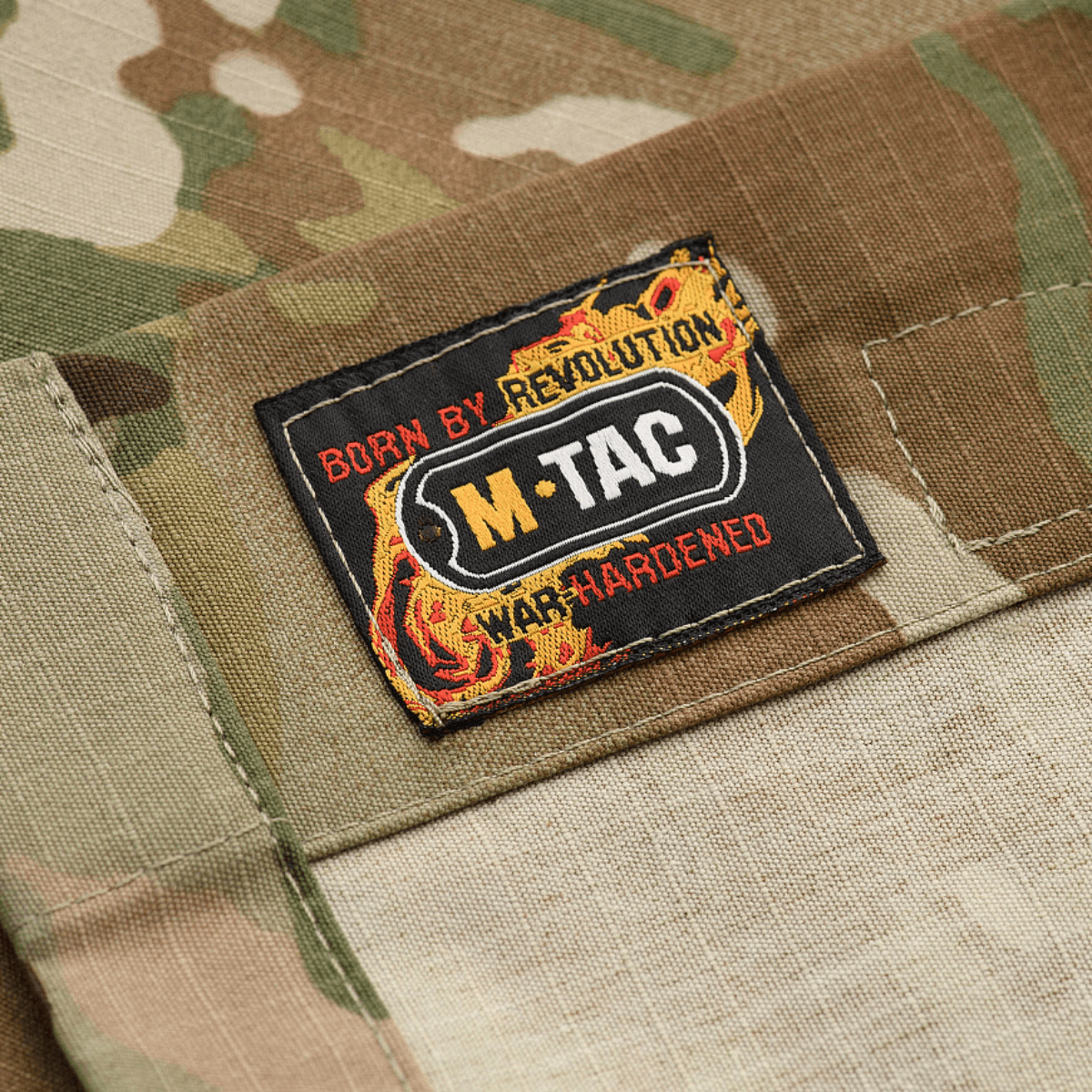 M-Tac Jakke Military Elite NYCO label on camouflage fabric, featuring "Born by Revolution, War Hardened" text and M-Tac logo.
