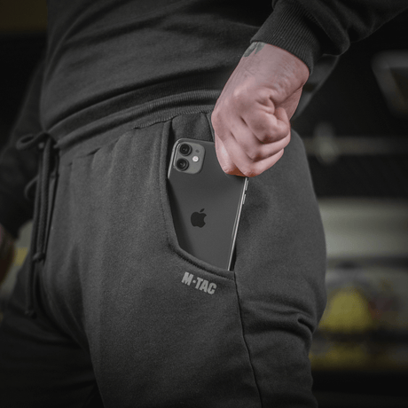 Person wearing M-Tac Cotton Classic pants with smartphone in side pocket, sportswear in 100% cotton offering comfort and freedom of movement.