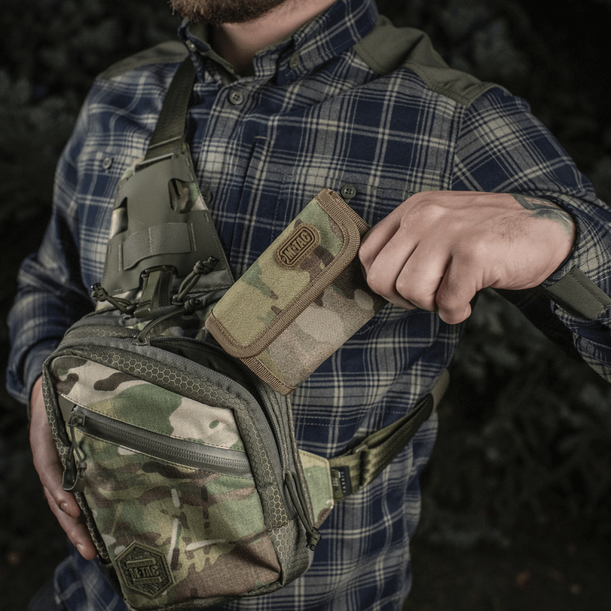 Mand holder M-Tac Pung Elite Gen.II i Cordura 500D ved siden af camouflage-rygsæk, udendørs miljø.
