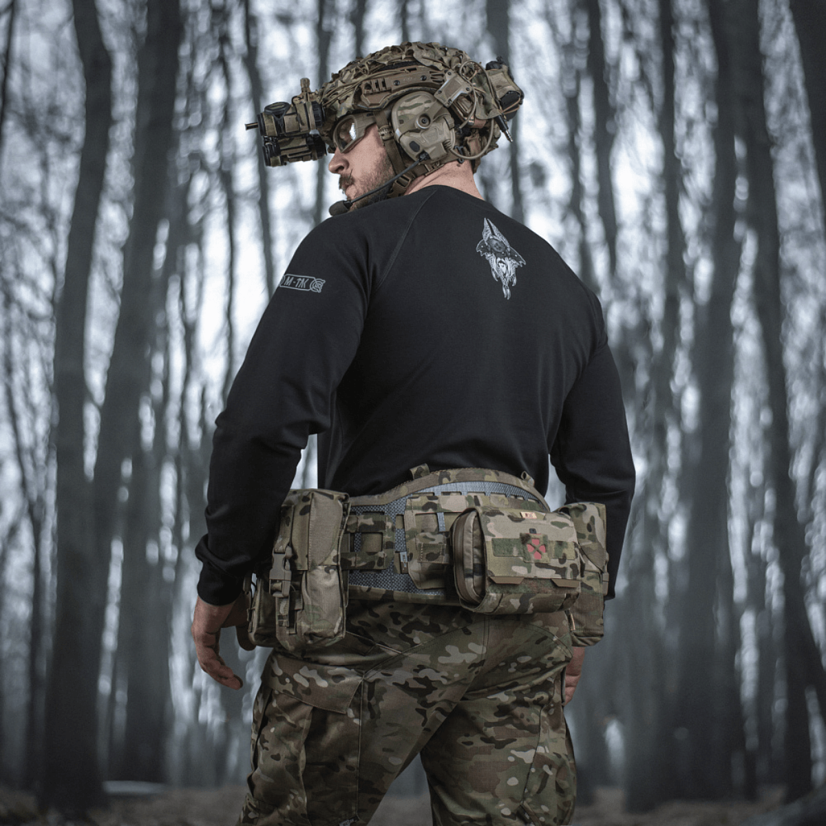 M-Tac Raglan Odin Mystery trøje med Odin-motiv, bæres af en person i taktisk udstyr i skovmiljø, fremhæver trøjens design og funktionalitet.