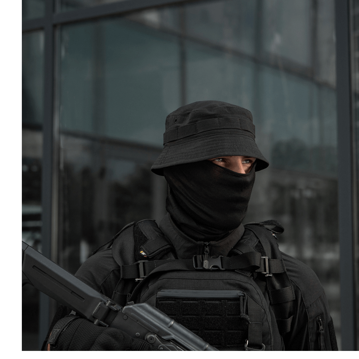 Person i sort taktisk udstyr og ansigtsovertræk holder våben foran en bygning.