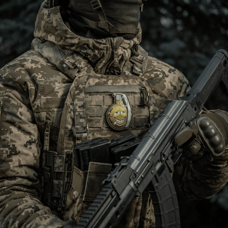 Soldier with camouflaged jacket wears M-Tac Patch M67 Grenade 3D PVC on vest, humorous emoji design, tactical military gear.