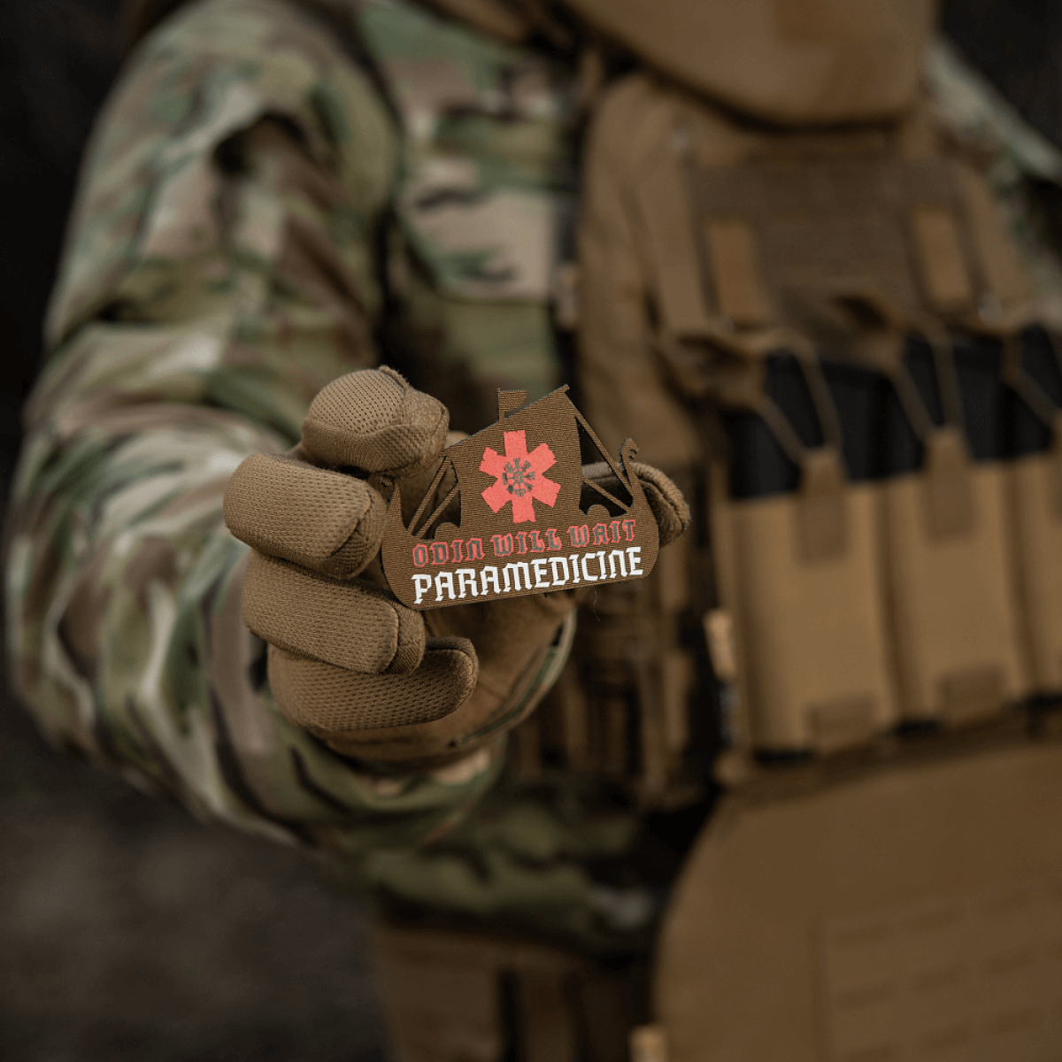 M-Tac Patch Viking Evac i soldatens hånd, viser Odin will wait paramedicine motiv. Camouflage jakke og taktisk vest baggrund.