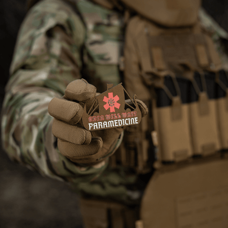 M-Tac Patch Viking Evac in a soldier's hand showing Odin Will Wait Paramedicine motif. Camouflage jacket and tactical vest background.