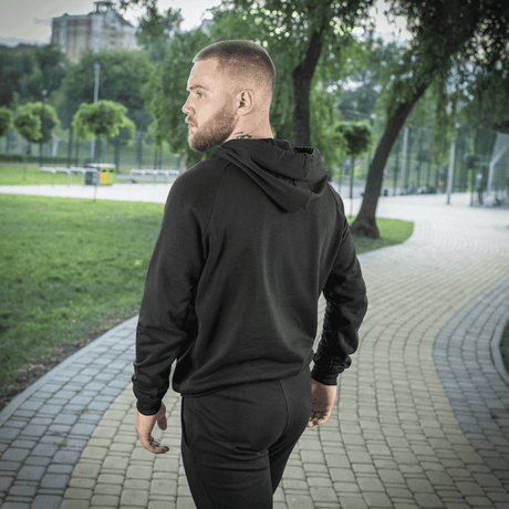 Mand iført M-Tac Hættetrøje Cotton Raglan i sort, gående i parkmiljø, understreger komfort og stil.