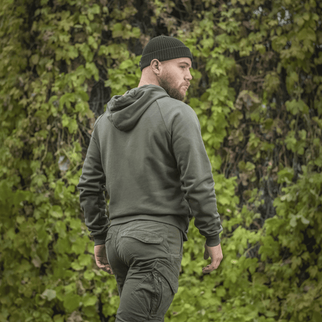Mand iført M-Tac Hættetrøje Cotton Raglan, står foran grønne blade, viser raglanærmer og hætte, komfortabel beklædning.