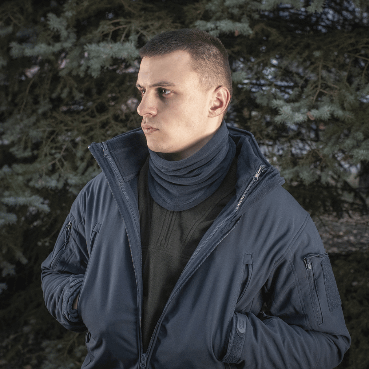Mand iført M-Tac Anatomisk Tube Scarf med Snor Polartec i en mørk jakke, stående foran træer, viser funktionelle anvendelser.