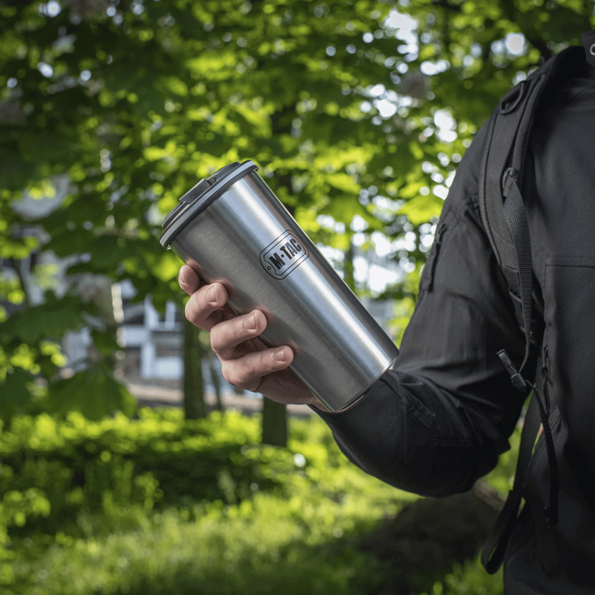 Person holder en M-Tac Termokop 450 ml med Ventil i rustfrit stål i en grøn have, ideel til varme og kolde drikke på farten.