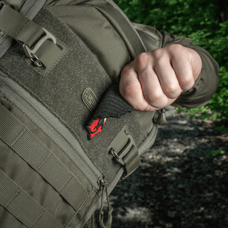 Person pulling an M-Tac Patch Husaria 3D PVC morale patch out of a tactical backpack, showing the winged hussar design against a green forest background.