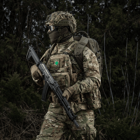 Soldier in military uniform with M-Tac Patch Lucky Card (Embroidered) attached to vest, symbol of luck and protection, camouflage in forest area.