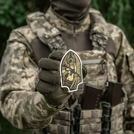 Soldier in camouflage uniform holding a M-Tac Valkyrie sticker depicting a war goddess with stylized helmet and M4 rifle.