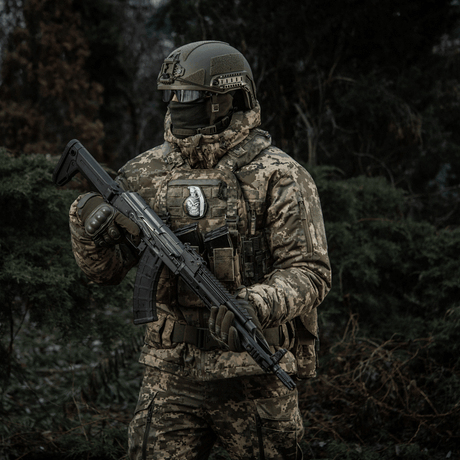 Soldier in camouflage with rifle in a forest, wearing M-Tac Patch M67 Grenade 3D PVC on a tactical vest