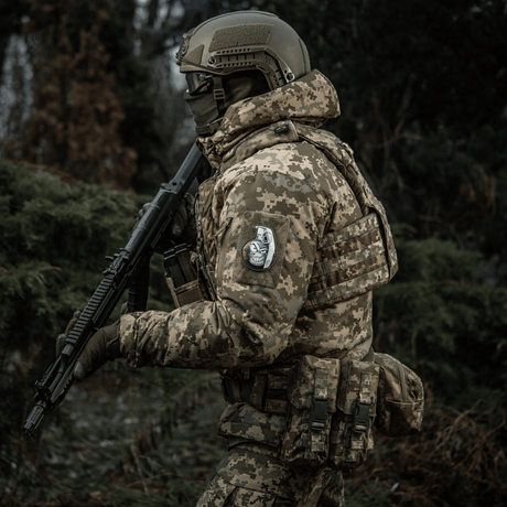 Soldier in camouflage uniform with M-Tac Patch M67 Grenade 3D PVC on sleeve, armed in forest.
