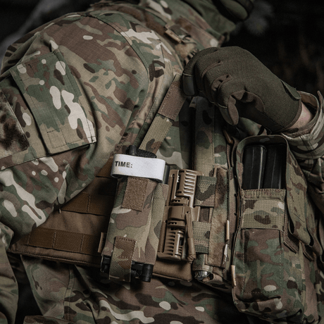 M-Tac Elastisk Tourniquet Pouch Molle Gen.III fastgjort til taktisk vest med tourniquet og ekstraudstyr i militær område.