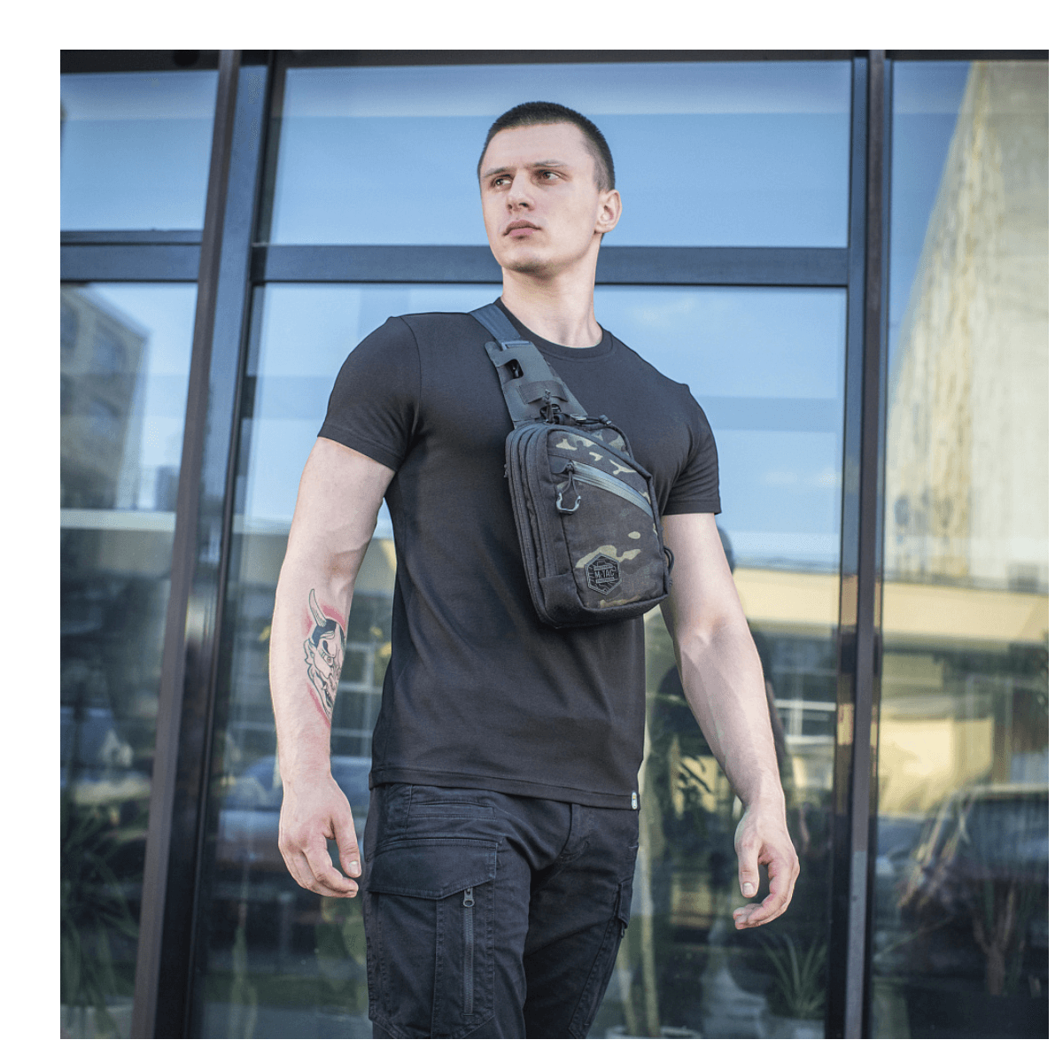 Mand iført M-Tac Sling Pistoltaske Elite Hex foran glasbygning, viser alsidighed og stil