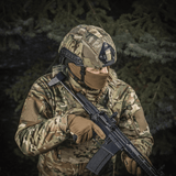 Person in camouflage gear wearing M‑Tac Hjelmcover Shroud Elite on helmet, holding a rifle in a forest environment.