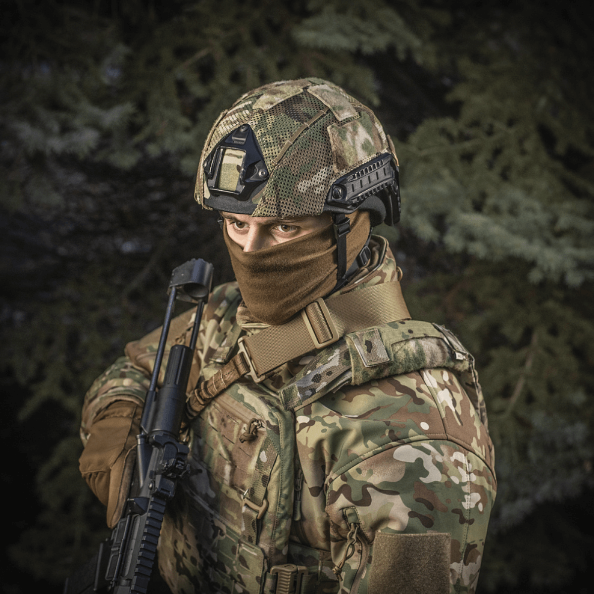 Person wearing M‑Tac Hjelmcover Shroud Elite in MULTICAM, holding rifle, displaying tactical gear outdoors.