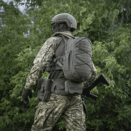 Soldat i camouflageuniform og taktisk udstyr i skovområde, bagfra