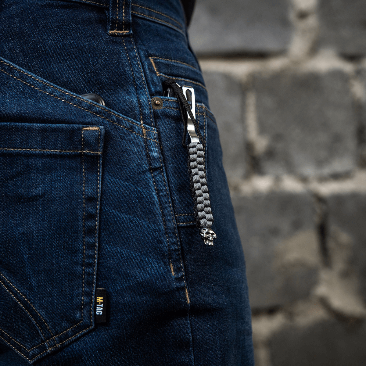 M-Tac Knife Lanyard Cuboid Skull med dekorativ perle, der hænger fra en knivlomme i blå jeans