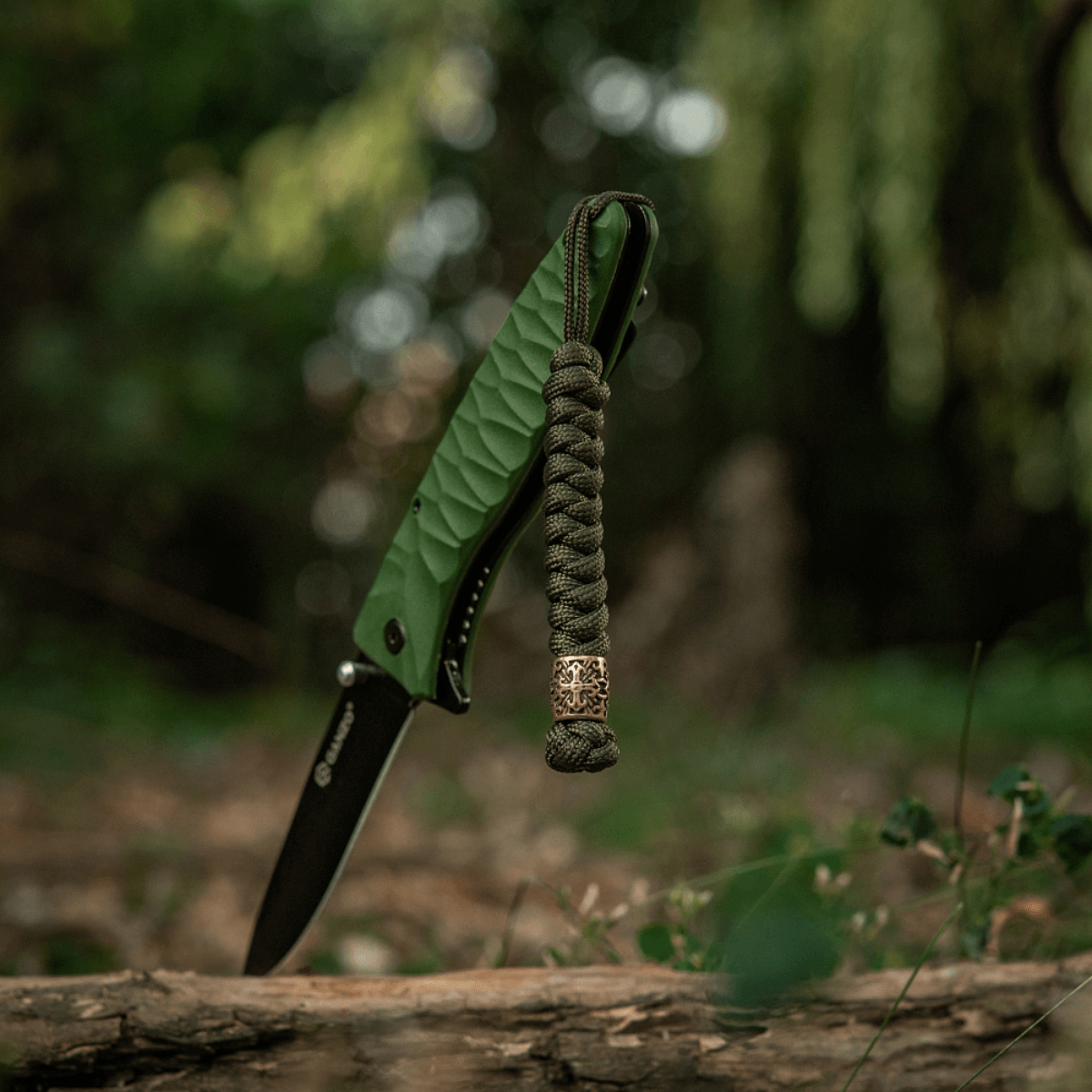 Grøn M-Tac Viper Lanyard Scandinavian paracord med flettede snore og viper-perle, hængende på skovbakke tæt på kniv i skoven.