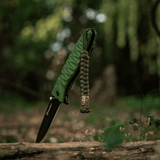 Grøn M-Tac Viper Lanyard Scandinavian paracord med flettede snore og viper-perle, hængende på skovbakke tæt på kniv i skoven.