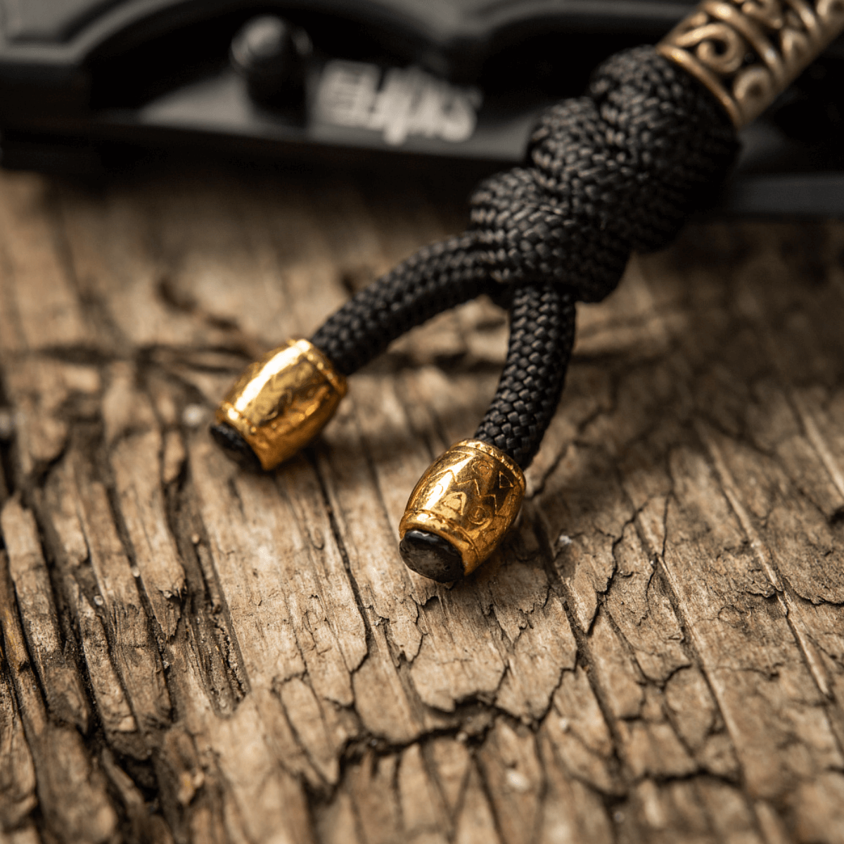 M-Tac Zeus Lanyard i sort flettet paracord med gyldne ender på træ overflade. Stilfuldt design til kniv eller udstyr.