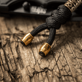 M-Tac Zeus Lanyard i sort flettet paracord med gyldne ender på træ overflade. Stilfuldt design til kniv eller udstyr.