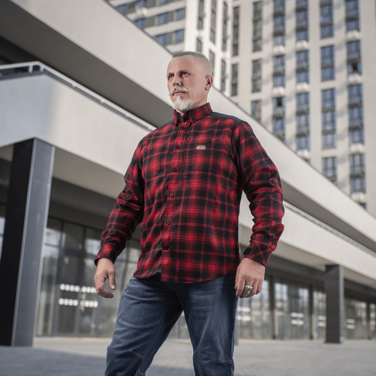 M-Tac Redneck Skjorte i rød-sort plaid på mand i urban baggrund, taktisk design, perfekt til arbejde og fritid.