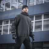 Mand iført M-Tac Alpha Gen.III Pro Vinterjakke med justerbar hætte, perfekt til koldt vejr og hverdagsbrug, baggrund med moderne bygning.