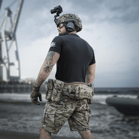 Soldat ved kyst i M-Tac T-Shirt Surf Club, camouflage shorts, hjelm og kameraudstyr, inspireret af ukrainske specialoperationer.