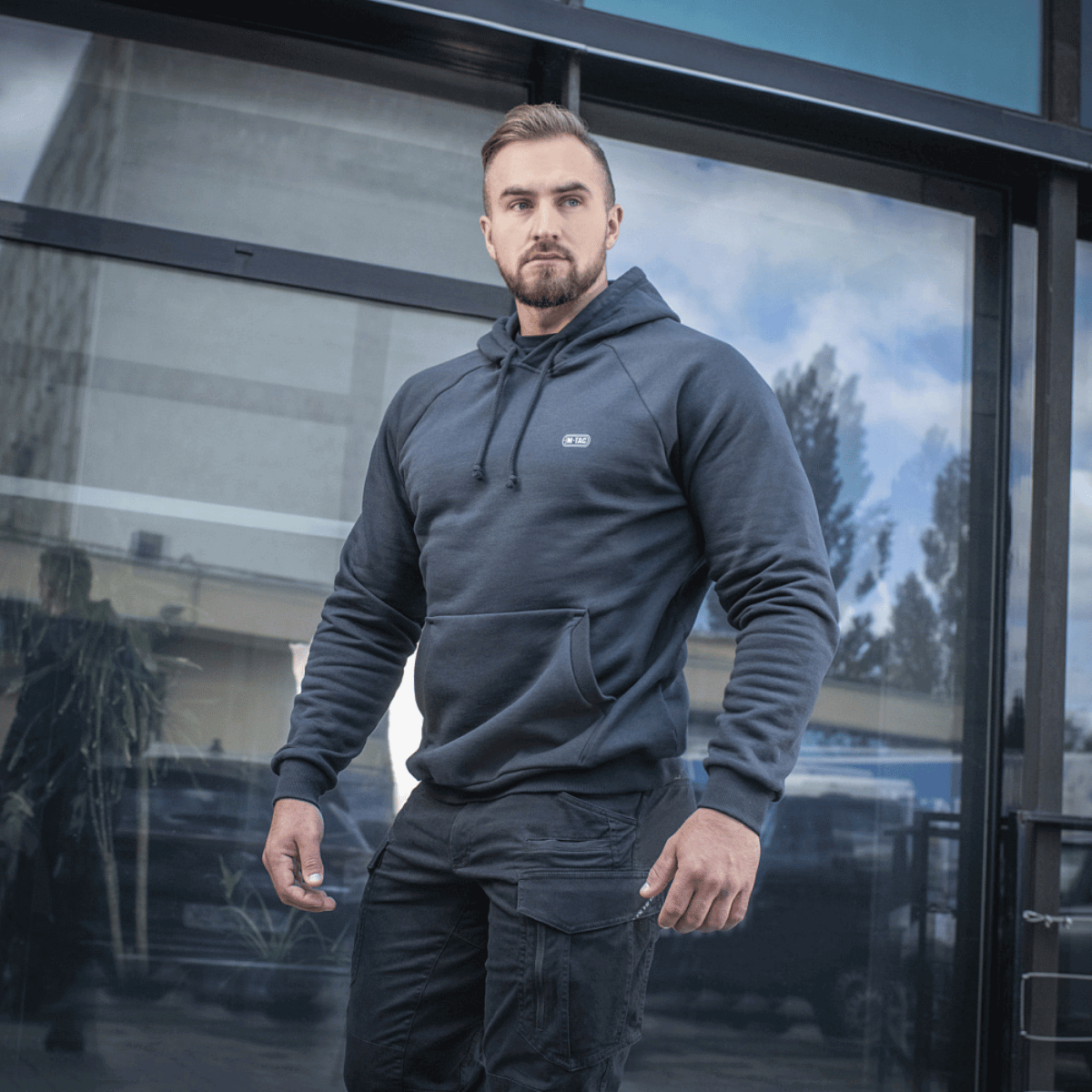 Mand iført M-Tac Hættetrøje Cotton Raglan med komfortabel pasform, egnet til både hverdags- og aktiv livsstil, foran glasbygning.