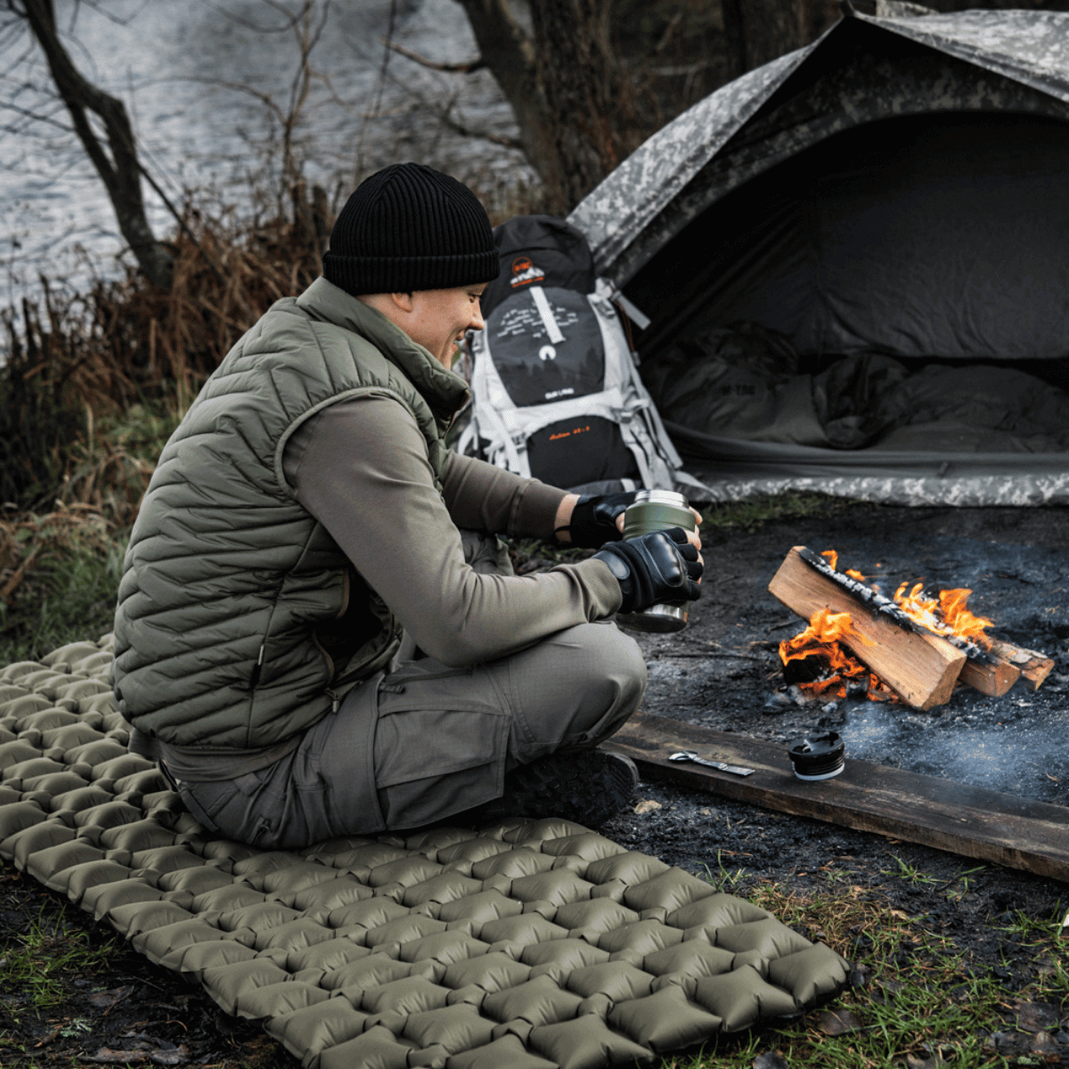 Mand sidder på M-Tac Oppustelig Liggeunderlag 195x60 tæt ved lejr, komfortabel camping ved bål