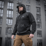 Mand i sort M-Tac Cotton Raglan Hard Hoodie med kængurulomme, stående udendørs foran en bygning, alsidig til hverdagsbrug.